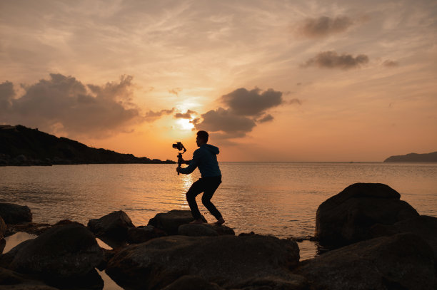 在海岸边的岩石上的人的剪影持有一个平衡环稳定器来拍摄日落的美丽风景图片下载