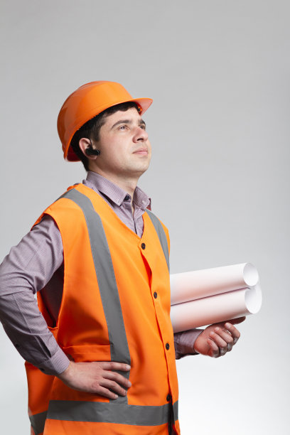 年轻的建筑工人戴着安全帽，在灰色的工作室背景上用手中的卷轴纸思考建筑项目图片下载