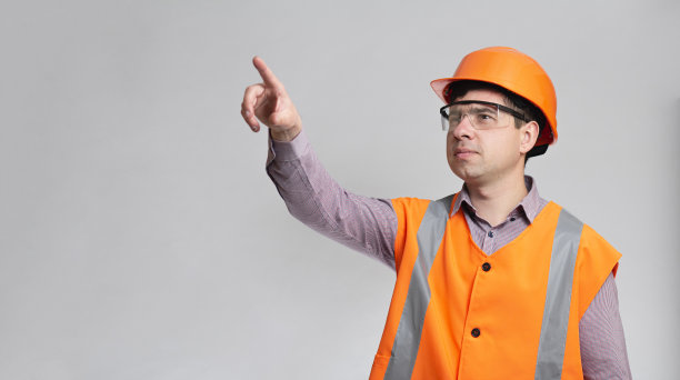 年轻的建筑工人穿着防护服在灰色的工作室背景显示与手指方向向前图片下载