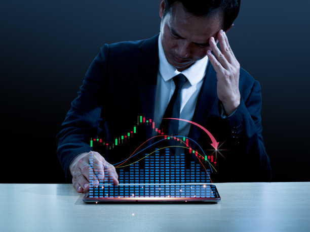 亚洲人在西装坐在他的桌子，左手在他的头，他做了悲伤的脸。右手放在桌子上。平板电脑一侧的屏幕上显示了一个股价大幅下跌的全息图。图片下载