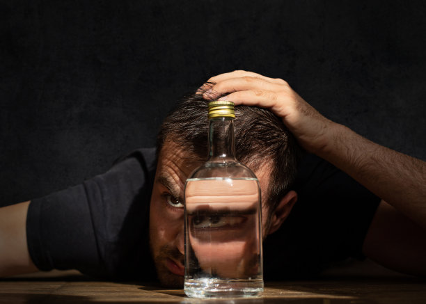 Alcololic上瘾。留着胡子的帅哥在黑暗的拷贝空间里拿着酒精饮料。桌子上放着一瓶伏特加的人图片下载