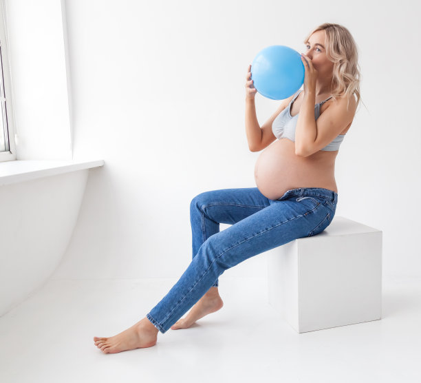 一位穿着蓝色牛仔裤、大肚子的美丽年轻孕妇拿着一个气球，表示即将诞生一个男孩。图片下载