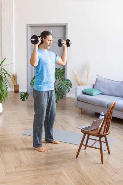 一个男人在家里用手提电脑举着哑铃锻炼的垂直照片。他听从在线教练的指导图片下载