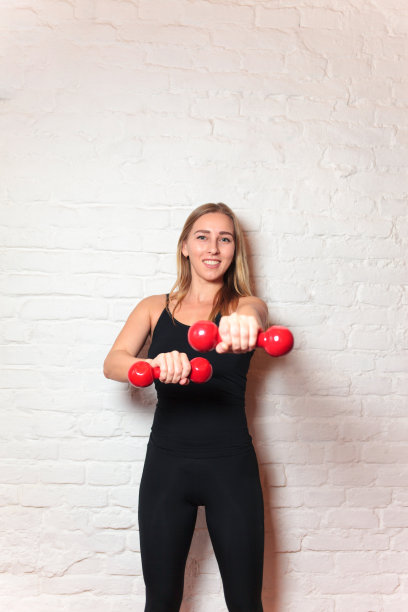 运动女孩穿着时尚的运动服拿着哑铃。灯光背景下的肌肉健身模型照片。运动与健康的生活方式图片下载