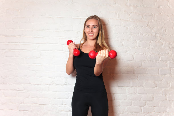 运动女孩穿着时尚的运动服拿着哑铃。灯光背景下的肌肉健身模型照片。运动与健康的生活方式图片下载