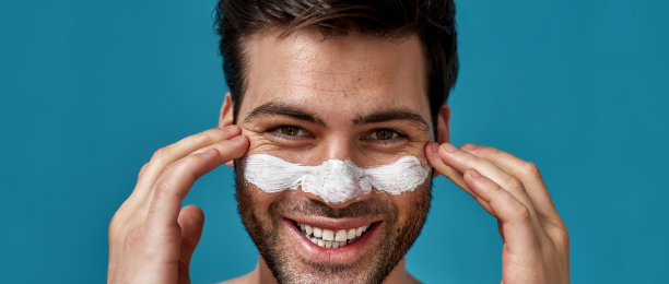 快乐的大胡子小伙，皮肤干净，用白色面膜清洁鼻子上的毛孔，触摸他的脸，在蓝色背景下对着镜头微笑图片下载
