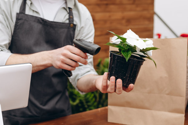 卖家扫描花盆条码的裁剪照片。鲜花递送服务和销售家庭盆栽，木橱窗。图片下载