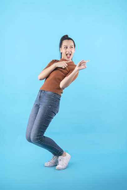 开朗的亚洲女性女孩表示拇指空白手势手指点宽的微笑张开的嘴穿着休闲的布料牛仔裤孤立的蓝色背景图片下载
