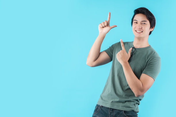 开朗的亚洲男性表示拇指空白手势手指点宽微笑张着嘴穿着休闲布料牛仔裤孤立的蓝色背景图片下载