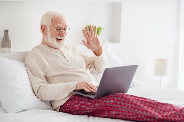 滑稽英俊的老人睡衣眼镜照片躺在床上挥舞手臂的现代装置在室内的家庭房间图片下载