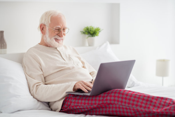 老人的照片，快乐，积极，微笑，坐在床上看，看，浏览，上网，笔记本电脑，打字，发邮件，穿睡衣回家图片下载