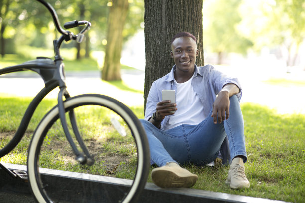 在城市公园里，快乐的黑色摩托车手坐在树旁拿着手机图片下载