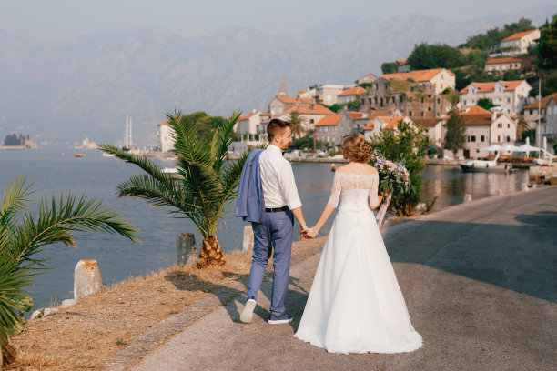 新郎和新娘手牵手站在佩拉斯特古城附近的码头上，新娘手里拿着一束花图片下载