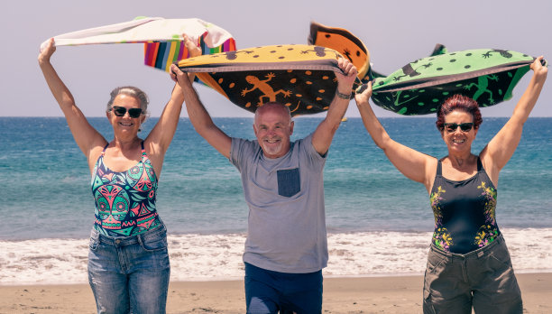 三个成熟的人站在沙滩上，在风中挥舞着毛巾，看着镜头。放松度假的概念图片下载