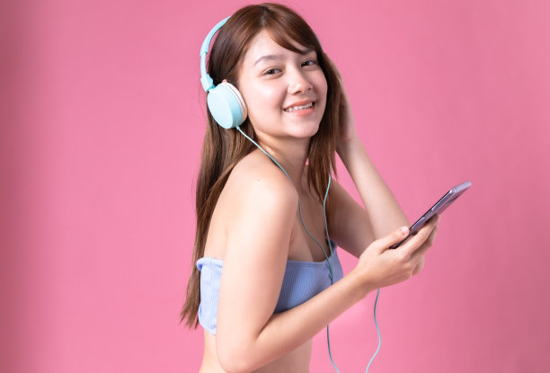 美丽，性感的亚洲女孩站在粉红色的背景。她穿着比基尼和微笑。暑假快乐旅行概念。混血女人听音乐。图片下载