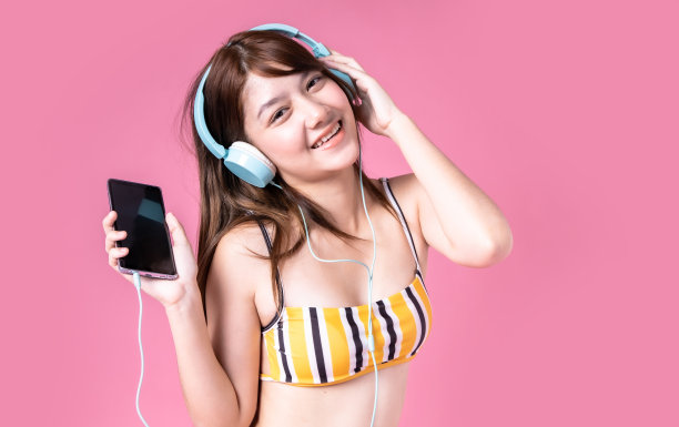 美丽，性感的亚洲女孩站在粉红色的背景。她穿着比基尼和微笑。暑假快乐旅行概念。混血女人听音乐。图片下载