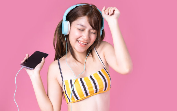 美丽，性感的亚洲女孩站在粉红色的背景。她穿着比基尼和微笑。暑假快乐旅行概念。混血女人听音乐。图片下载