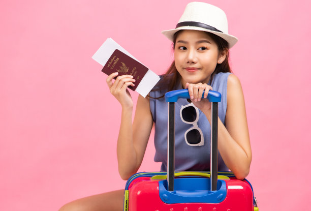 在粉红色的背景下，一个年轻的女人提着手提箱。漂亮可爱的梦幻女乘客的照片指着拷贝空间。图片下载