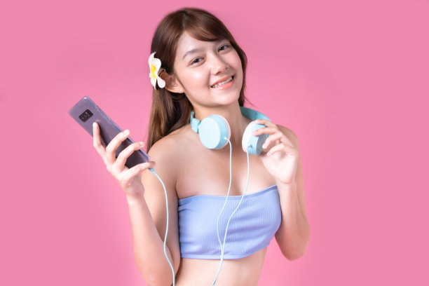 美丽，性感的亚洲女孩站在粉红色的背景。她穿着比基尼和微笑。暑假快乐旅行概念。混血女人听音乐。图片下载