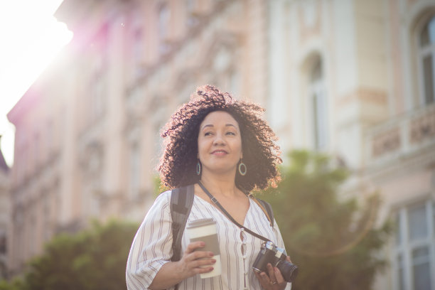 一个拿着相机站在街上喝咖啡的女人。图片下载