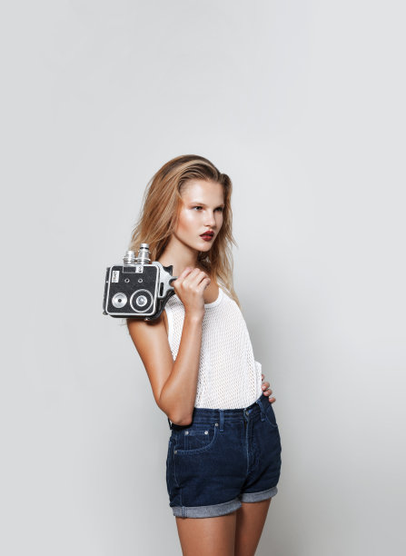 年轻女人的肖像金发碧眼摆姿势在工作室白色时尚衣服，看着别处，拿着一个复古相机在手里。图片下载