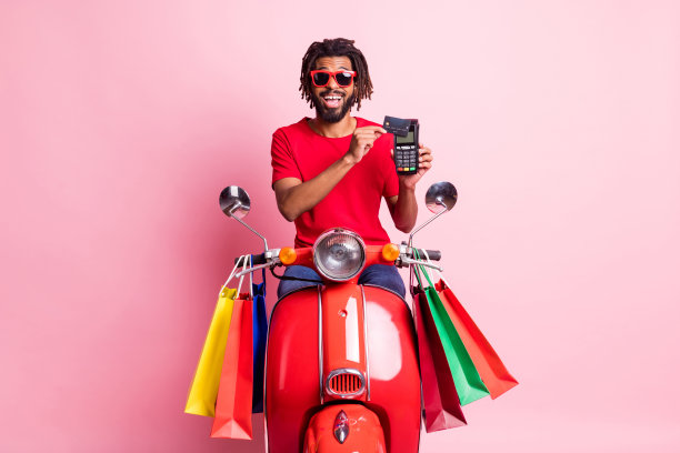 照片英俊的黑皮肤男子穿着红色t恤坐摩托车手持银行卡终端孤立粉红色背景图片下载