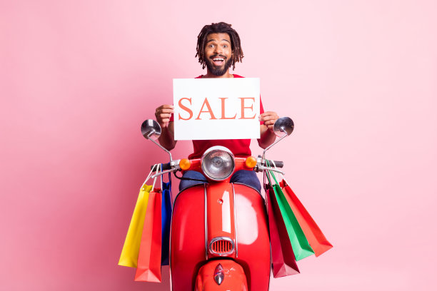 照片可爱英俊的黑皮肤男子穿着红色t恤，坐着摩托车抱着销售海报孤立的粉红色背景图片下载