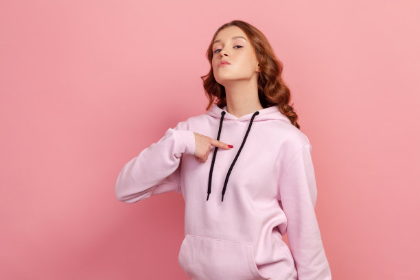 自私自利的褐发年轻女子在粉红色帽衫指向自己的手指，看着相机与傲慢和骄傲图片下载