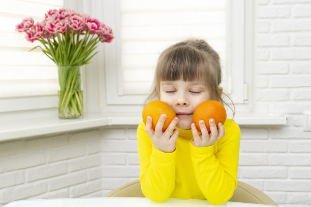 在白色的厨房里，一个小女孩坐在桌旁，闻着手中橘子的香气图片下载