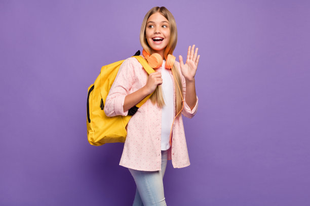 档案照片乐观的长头发漂亮的女孩挥手手戴背包耳机粉红色衬衫孤立在紫罗兰色的背景图片下载