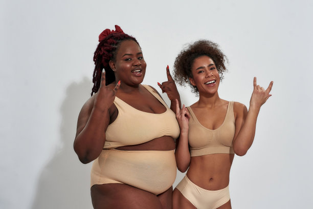 两个有趣的苗条和丰满的非裔美国女人穿着内衣在指示牌上做手势，在灰色的背景下孤立地一起摆姿势图片下载