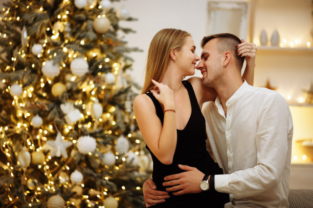 情侣们在圣诞树彩灯旁的沙发上亲吻、拥抱。新年的夜晚。圣诞节图片下载