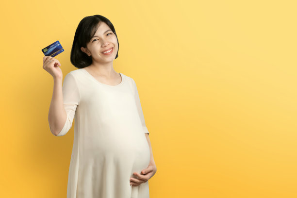 微笑的亚洲孕妇摸着肚子，手里拿着信用卡，在黄色的孤立背景下看着相机图片下载