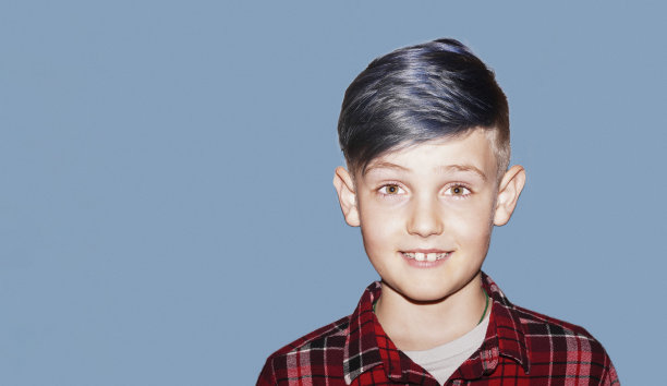 在灰色的背景上带着灿烂的微笑的快乐的小孩。金发少年与时尚流行的发型蓝色头发。一个男生站在那里看着摄像机。图片下载