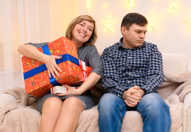 成人情侣的肖像，坐在家里的沙发上带着礼物，一个女人拿走一个男人的所有礼物，照片就像一个笑话，节日的灯挂在墙上图片下载