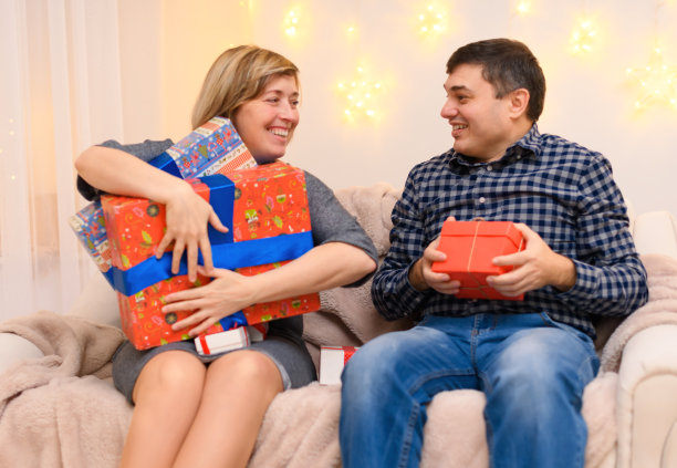 成人情侣的肖像，坐在家里的沙发上带着礼物，一个女人拿走一个男人的所有礼物，照片就像一个笑话，节日的灯挂在墙上图片下载