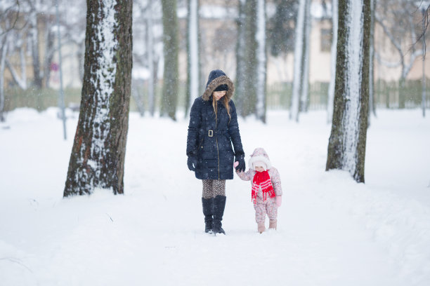 穿着工作服的小女儿和年轻的母亲走在公园的白雪上。在美丽的冬日里，一起度过时光。享受和平的漫步。前视图。图片下载
