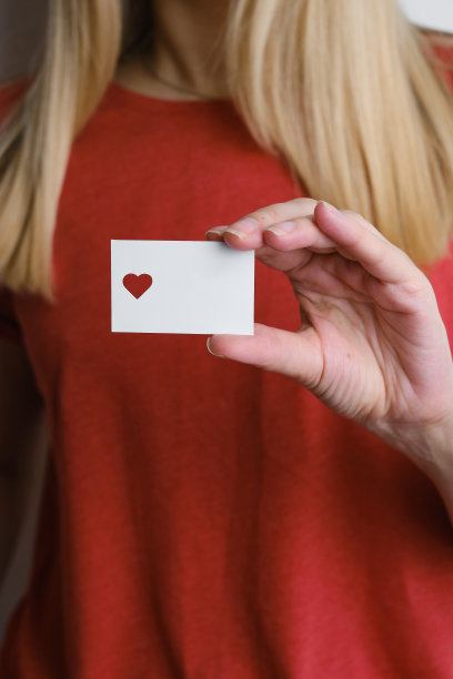 一个穿着红色t恤的女孩手里拿着一张白色卡片，上面有一个切出来的心形。特写的明信片。平的。情人节贺词。模型。可爱的信息或纸上的邀请图片下载