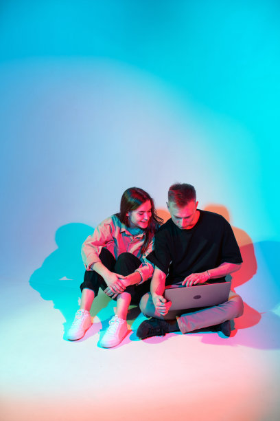 快乐的年轻时尚的夫妇用笔记本电脑浏览社交媒体，购物或在线玩。彩色的霓虹灯图片下载