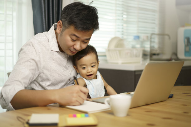 居家办公亚洲年轻的父亲写笔记本，把他的儿子放在膝盖上。看着笔记本。用笔记本电脑工作的企业家。一个男孩和爸爸坐在家里的厨房里。对冠状病毒大流行进行隔离。图片下载
