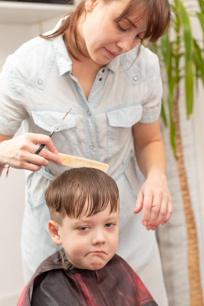 年轻漂亮的妈妈穿着浅蓝色的衣服，手里拿着梳子和剪刀在家给儿子理发。有选择性的重点图片下载