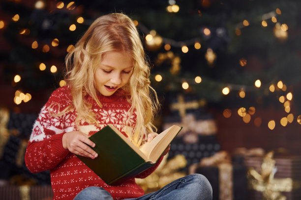 可爱的小女孩在圣诞节期间穿着红色的节日毛衣在室内看书素材图片