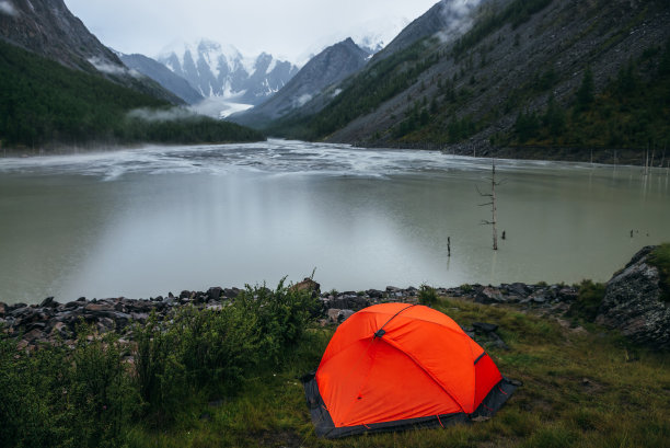 大气高山景观与橙色帐篷在岸边的绿色山湖和雪山在雨天。阴雨缭绕的山湖水面和低云的山谷景色图片下载
