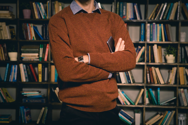 这个年轻人穿着毛衣和衬衫，手里拿着一部可以通过互联网打电话或下单的智能手机。它应该以一个放书的柜子为背景图片下载