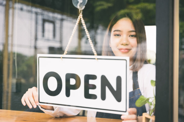 年轻的亚洲女孩翻转打开的标签欢迎顾客，并在早上打开咖啡馆。女亚洲咖啡师用镜子打开门口的咖啡馆。图片下载