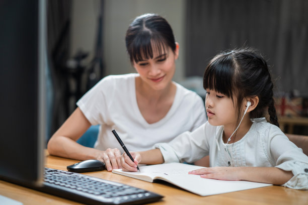 由于冠状病毒大流行，亚洲小女孩通过数字远程互联网会议从学校老师那里学习在线课程。孩子看着电脑，写着纸条，和妈妈坐在一起。图片下载
