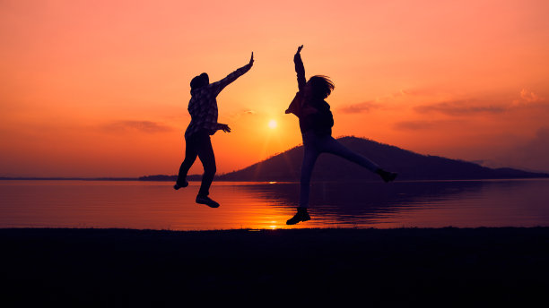 夕阳西下，两个人在湖边欢快跳跃的剪影。图片下载