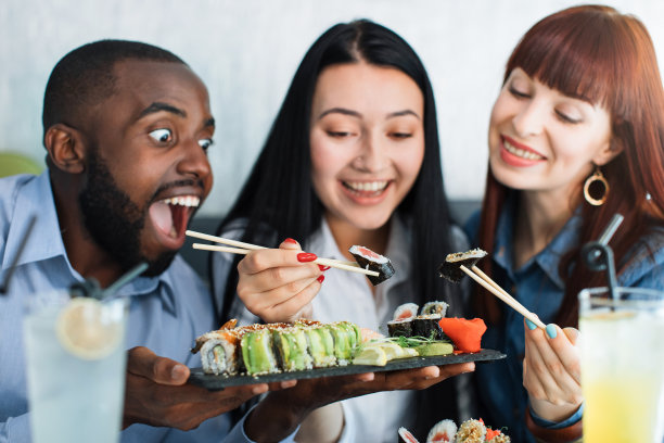 三个年轻的兴奋和饥饿的多民族人，吃着日本海鲜寿司卷在餐厅幸福和快乐。近距离的照片。朋友见面和寿司时间概念。图片下载