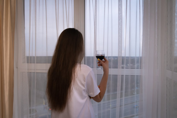 一位黑发长发、身穿白色特大号t恤的年轻女子站在窗前，手里拿着一杯红酒，望着远处的城市。背面照片图片下载
