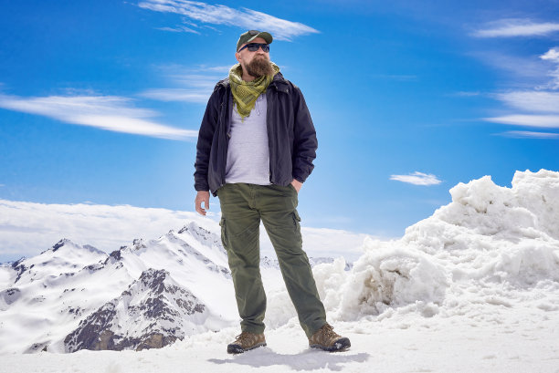 一名留着胡子、穿着游客服装的男子在白雪覆盖的群山背景下拍照。旅游冒险时尚图片下载